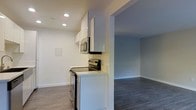 This single-floor space offers a clean, contemporary design with an open-concept layout that promotes a seamless flow between the kitchen and living areas. The kitchen is outfitted with modern white gloss upper cabinetry, providing ample storage while maintaining a bright, minimalist aesthetic. Stainless steel appliances, including an electric range and an over-the-range microwave, add a polished, functional touch. Light-toned countertops complement the crisp white walls and cabinetry, creating a cohesive, airy feel. Recessed lighting in the kitchen ensures excellent task illumination, while the overall neutral color palette keeps the space feeling open and inviting. Dark hardwood-style flooring runs throughout, grounding the interior with a rich contrast against the white walls and trim. A wide architectural opening connects the kitchen to the adjacent living area, enhancing the sense of spaciousness and making the layout ideal for both everyday living and entertaining. The living area benefits from the same consistent flooring and neutral tones, offering a versatile blank canvas. The space presents a well-maintained, move-in-ready interior with thoughtful design details throughout.