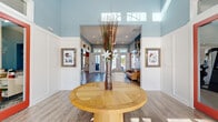Located at 50 Palma Vista Way in Saint Augustine, Florida, this single-floor clubhouse delivers a welcoming and thoughtfully designed community gathering space. The interior is defined by soaring ceilings and a bold dual-toned color palette — soft sky blue in the entry and lounge areas transitions into a vibrant terracotta orange in the social and kitchen zones — creating distinct yet cohesive atmospheres throughout.

The entry foyer greets visitors with a dramatic double-height ceiling, crisp white wainscoting, coastal-inspired wall art, and warm wood-look flooring that flows seamlessly throughout the space. A central round wood table anchors the foyer, setting an inviting tone from the moment you arrive.

The main lounge area features a rich blue accent wall with coffered paneling, comfortable seating, and large pendant lighting, ideal for relaxed gatherings. This transitions into a media lounge with a mounted flat-screen TV, built-in cabinetry, and additional flexible seating.

Floor-to-ceiling windows flood the space with abundant natural light and offer direct views to an outdoor terrace, where a glass door provides easy access. A well-appointed kitchenette with gray shaker cabinetry, open shelving, a decorative tile backsplash, stainless steel appliances, and a dining area completes the space, making it fully functional for events and everyday community use.