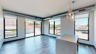 This single-floor unit showcases a contemporary urban design with industrial-inspired details seamlessly integrated throughout. The open-concept living and dining area is a standout, featuring wide-plank wood-style flooring, exposed silver ductwork along the ceiling, and an abundance of natural light streaming through large black-framed windows and a sliding glass door leading to a private balcony with outdoor views of the surrounding neighborhood.

The kitchen is thoughtfully designed with rich dark wood cabinetry, a classic white subway tile backsplash, granite countertops, and a center island with pendant lighting overhead. Stainless steel appliances, including a refrigerator, range, microwave, and dishwasher, complete the functional and stylish space. Track lighting and decorative glass pendant fixtures add warmth to the area.

A hallway connects the living spaces to the bedroom, which features plush carpeting, neutral walls, and a window offering additional natural light. Wood-style flooring continues through the corridors, tying the unit together cohesively. The overall ambiance is modern and polished, balancing industrial elements with warm, residential finishes.