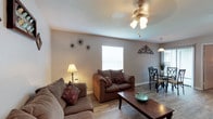 This single-story home is designed with a functional, open layout that connects the main living areas seamlessly. The living room features wood-look flooring, a ceiling fan with light fixture, and neutral-toned walls that create a warm, inviting atmosphere. Multiple windows allow natural light to filter in, and decorative wall accents add character to the space. The living area flows directly into the dining space, which is highlighted by a pendant light fixture overhead, a glass-top dining table, and a sliding door with vertical blinds that introduces additional natural light.

The kitchen is outfitted with dark cabinetry, granite-style countertops, a stainless steel sink, and black appliances, including a refrigerator and dishwasher. A pass-through opening connects the kitchen to the living area, enhancing the open feel.

The primary bedroom features plush carpeting, a window with blinds, and views into the dining area through an open doorway, along with a walk-in closet. A secondary bedroom with carpet flooring offers generous space suitable as a children's room or guest room. Two full bathrooms each include a tiled tub-shower combination and clean white finishes.