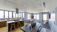 This single-floor space presents an open-plan layout that effortlessly integrates the kitchen, dining, and living areas into one cohesive and functional environment. The modern kitchen features a sleek island with a white countertop and built-in cooktop, complemented by a stainless steel range hood and bar seating with warm wooden stools — ideal for casual dining or entertaining. Adjacent to the kitchen, a rustic dark-wood dining table serves as a striking centerpiece, styled with decorative lanterns and offering ample seating. The living area is furnished with a comfortable neutral-toned sofa and coffee table, anchored beneath a clean white ceiling with recessed lighting and a cassette-style HVAC unit. Light hardwood flooring runs throughout, lending warmth and continuity across the space. Large windows on multiple walls flood the interior with abundant natural light and offer views of the surrounding cityscape and greenery. The cool grey-blue walls provide a calm, contemporary backdrop that ties the entire open-plan layout together. The overall ambiance is modern, airy, and welcoming — a well-appointed space designed for both comfort and style.