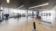 This expansive fitness center is thoughtfully designed to accommodate a full range of workout needs across two distinct zones. The main gym area features floor-to-ceiling glass curtain walls that flood the space with natural light while offering sweeping urban views, creating an energizing atmosphere for exercise. The herringbone-patterned wood-look flooring runs consistently throughout, adding a refined aesthetic that sets this facility apart from a typical gym environment. Structural columns are cleanly finished in white, contributing to the modern, polished design.

The main workout floor is comprehensively equipped with treadmills, ellipticals, stair climbers, rowing machines, stationary and cycling bikes, a full cable machine system, free weights, benches, and a punching bag, providing options for both cardiovascular training and strength work. A wall-mounted television and ample open floor space allow for flexibility in programming.

A separate studio room offers a distinct, intimate setting defined by a dramatic textured accent wall, dark ceiling, warm pendant lighting, full-length mirrors, and a ballet barre. This area is well-suited for yoga, stretching, or group fitness classes. Together, the two zones create a cohesive, upscale fitness environment with a strong emphasis on both function and design.