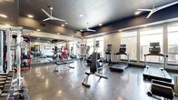 This fitness center offers a comprehensive workout facility spread across a single, open floor. The space is divided into two functional zones, each thoughtfully designed to support a full range of exercise activities. The main workout area features polished concrete flooring throughout, lending a sleek, modern aesthetic that is both durable and easy to maintain. A dark-painted ceiling with recessed lighting and large ceiling fans creates a dynamic atmosphere, while oversized windows along the perimeter invite natural light and provide views of the outdoors. Floor-to-ceiling mirrors amplify the sense of space and allow users to monitor their form during exercise. The cardio section is well-appointed with treadmills, ellipticals, stair climbers, and stationary bikes, many equipped with individual display screens. A dedicated strength training area includes a full dumbbell rack, weight benches, and cable machines. The secondary zone, distinguished by warm yellow accent walls and an industrial-style overhead door, serves as a functional training and spin studio, housing spin bikes, medicine balls, resistance bands, stability balls, and yoga mats. A glass-paneled entry door connects the space to the broader facility, completing this well-rounded fitness destination.