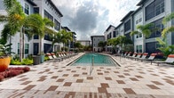 Located at 2410 Canopy Breeze Cir in Orlando, Florida, this outdoor amenity space presents a resort-inspired pool courtyard designed for relaxation and leisure. The centerpiece is a generously sized swimming pool with crystal-clear turquoise water, featuring a gradual entry staircase that invites easy access. The pool deck is finished in an elegant checkered pattern of light and earth-toned pavers, providing a visually striking and durable surface throughout the space.

Rows of sleek lounge chairs with vibrant orange accents line both sides of the pool, offering ample seating for sunbathing and outdoor relaxation. The courtyard is framed by lush tropical landscaping, including tall palm trees and colorful flowering plants, reinforcing the resort ambiance characteristic of Central Florida living.

The space is enclosed by modern multi-story residential buildings featuring a contemporary architectural style, with horizontal lap siding in cool gray and white tones, screened balconies, and large windows that overlook the courtyard. Ground-level units benefit from private screened patios with direct access to the landscaped surroundings.

The overall design creates an open-air environment that maximizes natural light and Florida's warm climate, blending functional outdoor living with refined aesthetic details throughout the communal area.