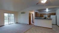 This single-floor space offers an open and airy layout that seamlessly connects the main living area to the kitchen. The living room is highlighted by vaulted ceilings that amplify the sense of space, complemented by a ceiling fan with a light fixture overhead. A wall-mounted mini-split air conditioning unit ensures year-round climate control, while a baseboard heater runs along the exterior wall beneath a sliding glass door that invites natural light into the room. Neutral-toned carpeting covers the living area floor, lending a warm and comfortable feel throughout.

The kitchen is positioned at an elevated counter level, creating a natural partition between the two spaces while maintaining an open sightline. It features warm wood cabinetry, dark countertops, and built-in appliances including a microwave and range, providing a functional and practical cooking environment. A hallway accessible from the main living space leads to additional rooms, with doors visible that suggest bedroom or closet access. The overall ambiance is clean, neutral, and functional, with a layout designed to maximize the utility of the single-floor footprint.