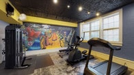 This fitness center offers a thoughtfully designed workout environment that balances functionality with distinctive character. The space is anchored by a striking, large-scale hand-painted mural spanning an entire wall, depicting a vivid scene of fantastical creatures and figures gathered around a campfire amid mountains and lush scenery — a lively artistic focal point that sets this gym apart from typical fitness facilities.

The flooring features a marbled, multi-toned epoxy surface that adds visual depth throughout the space. Overhead, a dark embossed tin ceiling with recessed and track lighting lends an industrial-meets-vintage aesthetic, creating a moody yet energizing atmosphere. Bold yellow accent walls and trim provide a bright contrast to the darker tones of the brick and ceiling.

Natural light filters in through multiple white-framed windows, illuminating the cardio equipment area, which includes several treadmills, an elliptical trainer, and a stationary cycling bike. A dedicated strength training area features a multi-functional cable machine and an adjustable weight bench, along with protective rubber floor mats. A wall-mounted television and fan offer added comfort during workouts. The fitness center is a compact yet complete facility, combining practical amenities with an artful, energetic atmosphere.