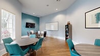 Located at 1767 Hermitage Blvd in Tallahassee, Florida, this professional meeting space combines functionality with contemporary design. The room is defined by a cool palette of soft gray and bold teal accent walls, complemented by warm hardwood flooring that runs throughout. Recessed lighting illuminates the space evenly, while large windows fitted with slatted blinds invite natural light and views of the surrounding greenery.

The space is thoughtfully organized into two distinct seating zones. A central conference table with built-in cable management ports is surrounded by a mix of ergonomic high-back chairs, making it ideal for formal meetings or collaborative work sessions. A wall-mounted flat-screen TV and a large whiteboard provide versatile presentation options. A secondary lounge-style seating area near the entrance features round tables and vibrant teal chairs, offering a more casual setting for smaller discussions or breakout conversations.

Additional details include a freestanding bookcase for storage and display, decorative ceramic vases, framed wall art, and a lush indoor plant that adds a touch of warmth. Glass-panel doors connect the room to adjacent common areas, enhancing the sense of openness. This space is well-suited for professional gatherings, team meetings, or collaborative sessions.
