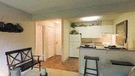 This single-floor space offers a thoughtfully designed open-concept layout that seamlessly connects the living area, kitchen, and hallway. The kitchen is outfitted with white cabinetry that extends to the upper walls, providing generous storage, while decorative plants atop the cabinets add a touch of greenery. A breakfast bar with a dark countertop and bar stool seating creates a casual dining option that bridges the kitchen and living space. White appliances, including a full-size refrigerator, complement the clean, bright aesthetic of the kitchen, which also features a sink and additional counter space. Wood-style flooring runs through the kitchen and hallway, offering both durability and a warm visual appeal, while carpeting in the living area provides comfort underfoot. The hallway, visible through an archway, leads to additional rooms and features a louvered bi-fold closet door, hinting at practical storage solutions. Soft overhead lighting throughout contributes to the warm, inviting ambiance of the space. The neutral wall tones and cohesive design elements create a comfortable and functional environment suitable for everyday living.