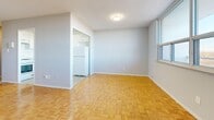 This thoughtfully laid-out 1-bedroom, 1-bathroom unit offers a practical and comfortable living arrangement across a single floor. The space is characterized by warm parquet hardwood flooring that runs throughout the main living areas and bedroom, lending a classic and timeless aesthetic.

The living room is a bright, open area bathed in natural light from large windows that offer elevated outdoor views, creating a spacious and airy feel. It connects seamlessly to the kitchen, which features white cabinetry — both upper and lower — marble-patterned countertops, a stainless steel sink, and light tile flooring, providing a clean and functional cooking environment.

The bedroom is a generously sized room with large windows that allow ample natural light and outdoor views. It includes a set of louvered sliding closet doors offering substantial built-in storage. The soft grey walls throughout the unit create a calm, neutral backdrop.

A narrow hallway with light wood-tone flooring and recessed ceiling lighting connects the main areas to the bathroom, which is finished with marble-effect wall tiles surrounding a full soaker bathtub and shower. The entrance provides a convenient transition into the unit. Overall, the space maintains a clean, modern aesthetic with a consistent and cohesive design language throughout.