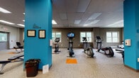 The fitness center at Wyncote House is a thoughtfully designed, single-floor facility offering a comprehensive range of exercise equipment and training zones. The space is organized into distinct areas, each serving a specific workout purpose, and is unified by a consistent design palette of warm taupe walls, teal accent columns, and vibrant orange floor markers that guide movement throughout the room.

The cardio zone is generously equipped with multiple treadmills, elliptical trainers, and stationary bikes, each positioned to take advantage of natural light streaming through several windows. Mounted televisions provide entertainment during workouts. The area features neutral carpeted flooring and a drop ceiling with recessed and panel lighting for consistent illumination.

A dedicated strength and free weights area includes a full rack of dumbbells, a weight bench, and a mirrored wall, accompanied by motivational text displayed on the walls. Stability balls of varying sizes are neatly stored on a rack nearby, supporting functional and flexibility training.

A separate open training room provides additional floor space for stretching or functional exercises, with clean white walls bearing motivational phrases such as "no excuses" and "motivation." Throughout the facility, the design balances practicality with an energetic atmosphere, making it a well-rounded fitness resource for residents of Wyncote House.