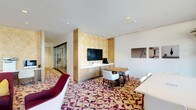 This single-floor suite showcases a thoughtfully designed open layout that combines a living area and dining space into one cohesive environment. The room is immediately distinguished by its bold geometric-patterned carpet in rich shades of magenta, cream, and gold, which anchors the space and adds a vibrant visual focal point underfoot.

The living area features a plush magenta accent chair, contributing a pop of color that complements the carpet's palette. Adjacent to this is a compact dining arrangement with a round wooden table and white upholstered chairs, ideal for in-room dining or casual meetings.

A built-in credenza in warm wood tones runs along the main wall, providing ample storage beneath a wall-mounted flat-screen television. The television is framed by a decorative panel featuring a subtle leaf-motif design in golden hues, adding an artistic architectural element. Recessed lighting in the white ceiling ensures bright, even illumination throughout.

A glass-paneled wall visible along the corridor adds transparency and a sense of depth, while framed artwork on the walls enhances the refined, hotel-suite aesthetic. The overall ambiance is polished, contemporary, and welcoming.