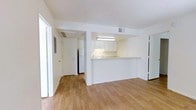 This single-floor space features a clean, minimalist design characterized by white walls and warm hardwood flooring that runs throughout the main living area and into the adjacent hallway. The open layout creates a seamless flow between the living space and the kitchen, which is accessible via a pass-through counter. This peninsula-style bar serves as both a functional workspace and a natural divider between the two areas without compromising the open feel of the space.

The kitchen is outfitted with white cabinetry and under-cabinet lighting that illuminates the countertops below. A full-size refrigerator is visible toward the rear of the kitchen, and the space includes standard cooking appliances. Overhead, a fluorescent light fixture provides ample task lighting throughout the kitchen area.

Ceiling-mounted HVAC vents are present throughout, ensuring climate control across the space. A hallway extending from the main living area leads to additional rooms, with a white door visible at the far end. The overall aesthetic is bright and neutral, with white surfaces and natural wood tones creating a light, airy atmosphere well-suited for a variety of uses.