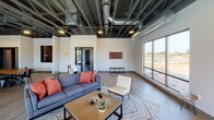 This single-floor amenity space showcases a compelling industrial-chic aesthetic throughout. The design is defined by dramatically exposed ceilings with dark-painted steel beams, structural elements, and visible ductwork, creating a loft-like atmosphere with generous overhead volume. Durable light wood-tone flooring runs seamlessly across the entire space, complementing the clean white walls.

The main lounge area is anchored by a striking floor-to-ceiling brick fireplace with a wall-mounted television, flanked by large sliding glass doors and expansive windows that invite abundant natural light and provide sweeping views of the surrounding landscape. Comfortable seating arrangements encourage relaxed gatherings in this bright, open environment.

A dedicated game room features a full-size pool table and casual dining-style table with industrial metal chairs, making it ideal for social recreation. Large windows in this area overlook an outdoor pool area, further enhancing the connection between indoor and outdoor amenities.

An additional lounge zone offers a more intimate seating arrangement with a sofa, accent chair, and coffee table, bathed in natural light from oversized windows with views of the open surroundings. A conference or meeting area with a wood table and black-framed glass-panel doors rounds out the functional offerings of this well-appointed, versatile space.