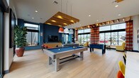 This single-floor amenity space is a thoughtfully designed communal hub that seamlessly blends recreation, relaxation, and social engagement. The open-plan layout is anchored by a central game room featuring a full-size billiards table with bold blue felt and a foosball table, both positioned beneath striking exposed wood beam ceiling elements that add warmth and architectural interest throughout the space.

Natural light floods the interior through expansive floor-to-ceiling windows and glass doors, offering views of the surrounding outdoor area, including a glimpse of a pool patio. Bold teal accent walls contrast beautifully with light hardwood flooring and crisp white ceilings, creating a lively yet inviting atmosphere. Colorful striped drapery panels and vibrant accent furnishings in red, yellow, and orange add a playful energy to the design.

A comfortable lounge area features plush sofas, armchairs, and a wall-mounted television, ideal for casual gatherings or unwinding. Adjacent seating nooks with yellow upholstered chairs and round ottomans provide quieter spots for conversation. The kitchenette area is outfitted with a white quartz island, multicolored bar stools, red pendant lighting, and a geometric tile backsplash. A welcoming reception lobby with modern ring pendant lights, tall indoor plants, and a sleek front desk completes the space.