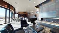 This striking single-floor space is defined by its bold contemporary design, combining luxurious materials and an open, flowing layout. The main living area serves as a visual centerpiece, anchored by a dramatic floor-to-ceiling fireplace clad in dark metal panels set against textured tile walls. A marble hearth surround adds elegance, while sleek black leather seating and a zebra-print area rug create a sophisticated, high-contrast aesthetic throughout. Recessed lighting illuminates the white ceiling, complementing the abundant natural light that streams through clerestory and oversized windows.

The kitchen area features an eye-catching curved wood ceiling, dark cabinetry, and a substantial curved marble-topped island surrounded by white upholstered barstools. Professional-grade appliances, subway tile backsplash, and pendant drum lighting complete the culinary space with both style and functionality.

A separate lounge area with floor-to-ceiling wood-paneled walls and a large garage-style window opens to an outdoor terrace, offering panoramic views and a serene spot for intimate seating. The entry corridor showcases rich horizontal wood paneling alongside a frosted glass door, setting a warm yet modern tone throughout the space.