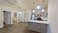 This thoughtfully designed one-bedroom unit offers a comfortable and functional single-floor layout with modern finishes throughout. The open-concept kitchen and living area serves as the heart of the home, featuring stainless steel appliances including a refrigerator, range, microwave, and dishwasher, complemented by soft gray cabinetry, white countertops, and a peninsula island with pendant lighting overhead. Durable wood-look flooring runs throughout the main living areas, adding warmth and continuity to the space. A glass-paneled exterior door in the living area allows natural light to fill the room. A dedicated in-unit laundry closet with a full-size washer and dryer provides added convenience. The generously sized bedroom features plush carpeting, a bright window with blinds, and direct access to a walk-in closet outfitted with dual hanging rods and ample shelving. The hallway connecting the living area to the bedroom is accented by a statement pendant light fixture, while a full bathroom with vanity and mirror is conveniently accessible. Neutral tones, clean lines, and well-placed lighting create a cohesive and inviting ambiance throughout the entire space.