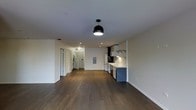 This single-floor space is designed with an open-concept layout that seamlessly integrates the living area and kitchen into one cohesive environment. Dark hardwood flooring runs throughout, providing a warm contrast against the crisp white walls and ceilings. The living area is generously proportioned, offering flexible arrangement possibilities.

The kitchen occupies one end of the space and is outfitted with white cabinetry, open wall-mounted shelving with black metal brackets, and a white tile backsplash. A stainless steel sink and white appliances, including a range and refrigerator, are neatly arranged along the kitchen wall. A dark lower cabinet unit adds a modern two-tone accent to the cabinetry design.

Lighting is provided through a combination of recessed ceiling fixtures and a bold black dome pendant light that serves as a focal point overhead. Toward the rear of the space, a hallway leads to additional areas, with a visible in-unit washer and a utility panel mounted on the wall. An entry door with a keypad lock is also visible, suggesting modern access control features throughout the unit.