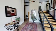 This single-floor space makes a strong first impression through its entryway, which combines industrial and refined design elements seamlessly. The foyer features tall ceilings with exposed overhead piping — a nod to industrial-style architecture — contrasted by warm, neutral-toned walls that create an inviting atmosphere. Large-format tile flooring grounds the space, complemented by a richly patterned area rug that adds warmth and visual interest underfoot. A wrought-iron console table with a glass top is positioned along one wall, styled with decorative greenery and accent pieces, contributing an elegant touch. A decorative framed mirror with ornate detailing is mounted on the far wall alongside a lush potted plant, enhancing the sense of depth and nature within the interior. A staircase with a modern metal railing and wood treads is visible to one side, suggesting multi-level access within the broader building. Natural light filters in through a window near the entryway's far end, brightening the corridor. Overall, the space balances raw architectural elements with carefully curated décor, resulting in a sophisticated and welcoming environment.