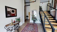 This single-floor space makes a strong first impression through its entryway, which combines industrial and refined design elements seamlessly. The foyer features tall ceilings with exposed overhead piping — a nod to industrial-style architecture — contrasted by warm, neutral-toned walls that create an inviting atmosphere. Large-format tile flooring grounds the space, complemented by a richly patterned area rug that adds warmth and visual interest underfoot. A wrought-iron console table with a glass top is positioned along one wall, styled with decorative greenery and accent pieces, contributing an elegant touch. A decorative framed mirror with ornate detailing is mounted on the far wall alongside a lush potted plant, enhancing the sense of depth and nature within the interior. A staircase with a modern metal railing and wood treads is visible to one side, suggesting multi-level access within the broader building. Natural light filters in through a window near the entryway's far end, brightening the corridor. Overall, the space balances raw architectural elements with carefully curated décor, resulting in a sophisticated and welcoming environment.