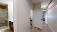Located at 205 E Grand River Ave in East Lansing, Michigan, this one-bedroom, one-bathroom unit offers a well-designed layout that maximizes every square foot. The open living and kitchen area sets a modern industrial tone, highlighted by exposed spiral ductwork running along the ceiling and light wood-style flooring that flows throughout the space. The kitchen is fully equipped with stainless steel appliances — including a range, over-the-range microwave, dishwasher, and refrigerator — complemented by warm wood cabinetry in two tones and clean white countertops. A dedicated dining nook with a compact table and chairs sits adjacent to the kitchen, while the living area accommodates seating and a coffee table beneath a large window fitted with roller shades that allow for natural light control. The bedroom, though cozy in scale, features a built-in bed frame with storage drawers and a sliding-door closet, providing practical storage solutions. Neutral gray walls and consistent light flooring maintain a cohesive aesthetic. The bathroom showcases a sleek, glass-enclosed walk-in shower with wood-grain tile surround, a dark wood vanity, and light tile flooring, lending a spa-like finish to the space.