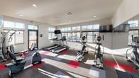 This fitness center is a thoughtfully designed, single-floor workout facility built to support a wide range of exercise routines. The space is flooded with natural light, thanks to an expansive bank of large windows along the perimeter walls, offering views of the surrounding exterior while creating a bright and energizing atmosphere. A glass-paneled entry door with transom windows further enhances the openness of the space.

The flooring features a patterned carpet tile layout in shades of gray and red, providing both durability and a visually dynamic aesthetic. Recessed lighting supplements the ample natural light, ensuring the space is well-lit at all times of day.

The cardio zone is well-stocked with multiple treadmills and elliptical trainers, each positioned to face the windows, allowing users to exercise with an outdoor view. Wall-mounted flat-screen televisions are strategically placed for convenient viewing during workouts.

A large floor-to-ceiling mirror on one wall visually expands the space and aids in form checking during exercise. The strength and functional training area includes a multi-station cable machine, a rack of dumbbells, a stability ball stand with various sized exercise balls, and additional free-weight accessories. Protective rubber floor mats are placed beneath equipment to safeguard both the flooring and users. The overall ambiance is clean, modern, and purpose-driven.