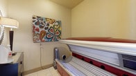 This single-floor space is designed as a dedicated tanning room, offering a private and functional environment for personal wellness use. The room is compact yet efficiently arranged, centered around a full-size tanning bed with a red and silver finish, equipped with multiple UV bulbs along both the upper and lower panels. A small personal fan is positioned nearby for comfort during use. The walls are finished in a warm, soft yellow tone that contributes to a welcoming ambiance, while light-colored tile flooring adds a clean, easy-to-maintain surface underfoot. A vibrant, large-scale canvas painting featuring bold floral and paisley patterns in red, teal, yellow, and green hangs on the wall, adding a decorative focal point and infusing the space with color and personality. The ceiling is smooth and unadorned, keeping the focus on the functional elements within the room. Overall, the space is straightforward and purpose-driven, combining practical tanning equipment with simple decorative touches to create a comfortable and visually appealing personal wellness area.
