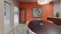 This clubhouse conference room is thoughtfully designed to balance functionality with contemporary style. The room's most striking feature is its bold terracotta-orange accent wall, which adds warmth and energy to the space. Anchoring the room is a large, dark oval conference table with integrated cable management ports, surrounded by patterned upholstered chairs in complementary orange and white hues. A wall-mounted flat-screen television is recessed into the neutral-toned wall opposite the accent wall, making it ideal for presentations and collaborative work sessions. Overhead, a sculptural white pendant light fixture adds an artistic touch, enhancing the room's refined aesthetic. The floor is finished with a textured, patterned carpet that adds visual depth while maintaining a professional atmosphere. A framed abstract artwork with geometric shapes in muted tones hangs prominently on the orange wall, reinforcing the curated design sensibility. A wood-trimmed door with a frosted glass panel provides privacy while maintaining a connection to the adjacent spaces. Glass partitions offer sightlines into a larger, lively lounge area beyond, which features exposed-beam ceilings, pendant lighting, and colorful seating — hinting at the broader, amenity-rich environment this conference room is a part of.