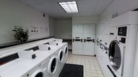 This single-floor laundry facility is designed with functionality and efficiency in mind, offering a clean and organized environment for shared use. The room features a row of front-loading commercial washing machines lined along one wall, each equipped with coin-operated or card-based payment controls for easy access. Opposite and adjacent, stacked dryer units maximize the use of vertical space, providing ample drying capacity alongside the washers. The space is finished with light-toned tile flooring that is easy to maintain and keep clean, while a drop-ceiling fitted with large fluorescent light panels ensures bright, even illumination throughout. A dark entry mat at the entrance helps keep the floor clean. The walls are neutral and uncluttered, with informational postings near the entrance providing usage instructions and guidelines. A folding or utility counter runs along one side of the room, offering a convenient surface for sorting and organizing laundry. The overall ambiance is practical and well-maintained, reflecting a thoughtfully arranged communal space built to serve the needs of its users efficiently.