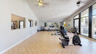 Located at 1030 S Dobson Rd in Mesa, Arizona, this fitness center offers a well-appointed exercise room designed to support a full range of workout routines. The space features a striking herringbone-patterned wood-look floor that spans the majority of the room, complemented by a dedicated rubber-mat zone for strength and functional training. The whitewashed wood-planked ceiling adds a warm, distinctive character to the interior, while recessed lighting and ceiling fans ensure a comfortable environment during workouts.

The room is equipped with a comprehensive selection of fitness machines, including treadmills, ellipticals, a recumbent bike, an upright cycle, and a multi-function cable resistance machine. Free weights, kettlebells, medicine balls, and a Matrix adjustable bench round out the strength training offerings. A large wall mirror on one end enhances the sense of depth and allows users to monitor their form.

Natural light flows in through large windows and a glass door, offering views of the surrounding landscaping and keeping the atmosphere bright and inviting. A wall-mounted television adds convenience for entertainment during cardio sessions. The clean, neutral color palette of white and light grey walls contributes to an open, energizing ambiance well-suited for daily fitness use.