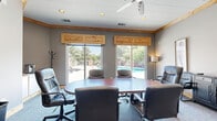 Located at 2727 Nelson Rd in Longmont, Colorado, this conference room offers a functional and comfortable meeting environment with a distinctive blend of rustic and professional design elements. The room is centered around a large oval conference table surrounded by leather executive chairs, providing ample seating for group gatherings or business meetings.

Natural light pours in through two expansive sliding glass doors fitted with warm knotty pine valances, which frame views of an outdoor pool area and surrounding mature trees. The same natural wood detailing is carried through the crown molding and entryway double doors, lending the space a cohesive, lodge-inspired aesthetic. Neutral gray walls complement the carpeted flooring underfoot.

A wall-mounted flat-screen television serves as a focal point for presentations, while a freestanding coat rack and a corner cabinet with a countertop—equipped with a single-serve coffee maker—add practical convenience. Additional seating is available along the perimeter, alongside a dark credenza and tasteful framed artwork. Recessed lighting and a ceiling fan ensure the room remains well-lit and comfortable throughout the day. The overall ambiance is professional yet inviting, making this space well-suited for productive meetings in a relaxed Colorado setting.