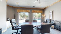 Located at 2727 Nelson Rd in Longmont, Colorado, this conference room offers a functional and comfortable meeting environment with a distinctive blend of rustic and professional design elements. The room is centered around a large oval conference table surrounded by leather executive chairs, providing ample seating for group gatherings or business meetings.

Natural light pours in through two expansive sliding glass doors fitted with warm knotty pine valances, which frame views of an outdoor pool area and surrounding mature trees. The same natural wood detailing is carried through the crown molding and entryway double doors, lending the space a cohesive, lodge-inspired aesthetic. Neutral gray walls complement the carpeted flooring underfoot.

A wall-mounted flat-screen television serves as a focal point for presentations, while a freestanding coat rack and a corner cabinet with a countertop—equipped with a single-serve coffee maker—add practical convenience. Additional seating is available along the perimeter, alongside a dark credenza and tasteful framed artwork. Recessed lighting and a ceiling fan ensure the room remains well-lit and comfortable throughout the day. The overall ambiance is professional yet inviting, making this space well-suited for productive meetings in a relaxed Colorado setting.