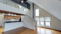 This two-floor loft-style space showcases an impressive open-concept design defined by dramatic vaulted ceilings that rise to striking heights on the main level. Warm hardwood flooring flows consistently throughout both floors, complementing the clean white walls and lending a cohesive, inviting atmosphere. Tall vertical windows on the main floor flood the living area with abundant natural light, while a sleek metal-railed staircase with a carpeted tread serves as both a functional and architectural focal point, connecting the two levels.

The kitchen is a well-equipped galley-style space featuring rich wood cabinetry, dark granite countertops with attractive speckled detailing, a stainless steel sink, and black appliances. A rustic wood beam accent above the kitchen area adds warmth and character, contrasting beautifully with the industrial-style exposed ductwork visible at the upper level. The kitchen opens toward the main living area, which also benefits from an overhead view of the upper loft through a metal railing.

Upstairs, the loft level offers a generously sized room with angled ceilings following the roofline, dual skylights that invite natural light, and a ceiling fan for added comfort. The bathroom features a clean, all-white tiled shower-tub combination with chrome fixtures, maintaining the space's bright and modern aesthetic throughout.