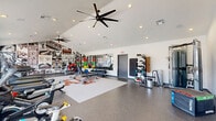Located at 1800 Holleman Dr in College Station, TX, this fitness center offers a spacious, single-floor layout designed to support a full range of workout routines. The facility is thoughtfully organized into distinct exercise zones, accommodating cardio, strength training, and cycling activities with ease.

The cardio area features multiple treadmills and stair climbers positioned along a wall of large windows, providing abundant natural light and direct views of the outdoor pool area and surrounding property. Nearby, a dedicated cycling zone includes stationary spin bikes, also benefiting from floor-to-ceiling glass panels that open the space visually to the outdoors.

The strength and free weights section is anchored by a bold Texas A&M-themed floor mat and flanked by a dramatic full-wall mural showcasing Aggie athletics, reinforcing the spirited collegiate atmosphere. A comprehensive dumbbell rack, adjustable benches, and additional strength equipment complete this zone. Wall-mounted storage organizes accessories and resistance tools neatly.

The interior features soaring vaulted ceilings fitted with oversized ceiling fans and recessed lighting, maintaining airflow and a bright, energetic ambiance. Durable speckled flooring runs throughout, practical for high-traffic athletic use. The overall design is modern, clean, and motivating—purpose-built for an active lifestyle.
