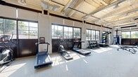 Located at 321 Ellis St in New Britain, Connecticut, this fitness center combines industrial character with modern amenities across a generously sized single-floor layout. The main exercise area showcases exposed wood-beam ceilings, white-painted brick walls, and visible ductwork, lending the space a loft-like, warehouse-inspired aesthetic. A long row of large, dark-framed windows lines one wall, flooding the floor with abundant natural light while offering views of the outdoor surroundings. The durable speckled rubber flooring throughout provides both safety and style suited for high-activity use. The cardio zone is well-appointed with multiple treadmills, an elliptical trainer, and a stationary bike, while the strength area features a multi-functional cable machine, a power rack with resistance bands, and a full dumbbell rack. Wall-length mirrors in the strength section enhance the sense of space and support proper form. A separate auxiliary room, distinguished by a vibrant, artistic mural and soft rubber flooring, provides a dedicated area for flexibility, balance, and functional training, complete with stability balls and a BOSU trainer. An additional area with built-in wooden shelving offers organized storage. The space also connects to an adjacent lounge area visible through glass-paneled doors, adding a social dimension to the facility.
