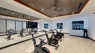 This single-floor fitness studio is thoughtfully designed to support both cycling workouts and broader exercise activities. The cycling area is equipped with multiple ICG stationary bikes arranged on light wood-look flooring, providing ample space between each station for comfortable use. A full wall of floor-to-ceiling mirrors runs along one side, visually expanding the room while allowing users to monitor their form during sessions. The ceiling is a standout architectural feature, showcasing a decorative textured tile pattern framed by warm wood trim, complemented by recessed lighting and a ceiling fan that ensures proper air circulation. A wall-mounted flat-screen display adds a multimedia element to the cycling area. Framed athletic photography adorns the walls, contributing to a motivating, professional atmosphere. A rack of dumbbells and step platforms are also present, adding versatility to the space. Large glass sliding doors and full-height glass partitions connect the cycling studio to an adjacent strength training area, promoting an open, cohesive feel throughout the facility. The transparent boundaries allow natural light to flow between zones while maintaining acoustic separation. The overall ambiance is clean, modern, and well-appointed.