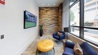 This single-floor space blends contemporary design with warm, textured materials to create a polished and inviting environment. The entry is defined by sleek glass-and-stainless-steel doors that open onto large-format stone tile flooring, offering a refined first impression. Inside, a striking reclaimed wood accent wall serves as a bold focal point, its rich, multi-toned planks adding warmth and organic texture against the clean white walls and modern finishes. The lounge area is thoughtfully appointed with deep navy armchairs, striped accent pillows, and a metallic side table, all arranged to take advantage of the expansive floor-to-ceiling windows that flood the space with natural light and frame views of the surrounding urban streetscape. A wall-mounted flat-screen television and a golden leather ottoman round out the lounge, making it a comfortable and functional gathering spot. Adjacent to the lounge, a dedicated business center features a long built-in desk accommodating multiple workstations, complete with computers and a printer. Sheer drapery softens the natural light, while abstract artwork adorns the walls, adding a creative touch. Patterned carpet tiles flow throughout, tying the varied zones together cohesively. The overall ambiance is sophisticated yet approachable, ideal for both focused work and relaxed socializing.