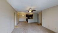 This single-floor residence offers a welcoming open-concept design that integrates the living, dining, and kitchen areas into one cohesive, functional space. The living area is finished with plush wall-to-wall carpeting that adds warmth underfoot, while a ceiling fan with an integrated light fixture serves as a central element, providing both comfort and illumination. A pendant light over the dining area adds a subtle decorative touch and helps define the space within the open layout. The kitchen is positioned at the heart of the home, anchored by a raised breakfast bar that creates a natural divide between the kitchen and living spaces. Dark cabinetry provides strong contrast against lighter surfaces, and the kitchen is equipped with essential appliances including a refrigerator, range, and microwave. Track lighting above the kitchen workspace ensures ample task lighting. Neutral-toned walls throughout create a clean, versatile backdrop adaptable to a variety of interior styles. A door visible from the main living area suggests convenient access to additional rooms or utility spaces. The overall ambiance is practical and comfortable, well-suited for everyday living.