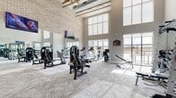This fitness center is a thoughtfully designed workout facility that combines architectural appeal with a comprehensive range of exercise equipment. The space is defined by its dramatic double-height ceilings with exposed wooden truss detailing, lending an open, airy character that sets it apart from a typical gym environment. Textured brick accent walls contrast with smooth, neutral-toned drywall, creating a modern yet warm aesthetic throughout.

Large, multi-pane windows line the perimeter, flooding the interior with abundant natural light and offering views of the surrounding landscape. Full-length mirrors along one wall visually expand the space while serving a functional purpose for form-checking during workouts. Patterned, low-pile carpet covers the flooring, providing comfort underfoot across the entire facility.

The equipment layout is well-organized and diverse. A row of treadmills and elliptical trainers supports cardio training, while a selection of selectorized strength machines, a Smith machine with free weights, and a full dumbbell rack cater to strength and resistance training. Recumbent and upright stationary bikes add additional cardio options. Stability balls and a mat area provide space for functional and flexibility training. Wall-mounted flat-screen televisions are positioned throughout, enhancing the workout experience. The facility is designed to support a wide range of fitness goals in a bright, spacious environment.