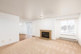 This single-floor residence offers a well-organized layout with distinct living, dining, kitchen, and bedroom areas. The spacious living room serves as the heart of the home, anchored by a tile-surround fireplace with glass doors and complemented by plush wall-to-wall carpeting. Natural light filters in through a window with blinds, while a sliding glass door provides convenient access to an outdoor area. Adjacent to the living room, the dining area transitions to warm hardwood-style flooring and features a pendant light fixture overhead, with a second sliding glass door drawing in ample natural light. The kitchen is efficiently designed with white cabinetry, dark granite-style countertops, and hardwood-style flooring. It is equipped with white appliances including a refrigerator, electric range, and built-in microwave. A peninsula with a sink adds both prep space and a casual breakfast bar. A window above the counter brings in natural light and outdoor views. The bedroom is a quiet, carpeted room with clean white walls offering a blank canvas for personalization. The bathroom includes a full tub-shower combo, a toilet, hardwood-style flooring, and a vanity with matching granite-style countertops, maintaining a cohesive aesthetic throughout the home.
