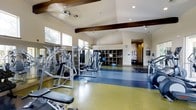 This fitness center is a thoughtfully designed, single-floor facility that combines functionality with an inviting architectural aesthetic. The space is characterized by soaring vaulted ceilings accented with prominent exposed wooden beams, lending a warm, structured visual element to the otherwise clean, neutral interior. A ceiling fan aids in air circulation, contributing to a comfortable workout environment.

The flooring is divided into distinct zones, with a blue rubberized surface designated for strength training equipment and a yellow-green area suited for open-floor exercises or stretching. A comprehensive selection of cable and weight machines is arranged throughout the main training floor, providing options for a full-body workout routine.

Along one wall, a built-in shelving unit offers organized storage for exercise accessories such as stability balls and other equipment. Large windows along the upper walls allow natural light to filter into the space, enhancing the overall ambiance. Mirrored wall panels further amplify the sense of openness. A secondary area visible in the background appears to provide additional functional space, extending the overall utility of this well-appointed fitness facility.