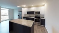 This single-floor residence showcases a clean, modern aesthetic defined by a cohesive blend of dark and light tones throughout. The open-concept kitchen serves as a focal point, featuring sleek two-tone cabinetry with dark lower cabinets and white upper cabinets, granite countertops, and a full suite of stainless steel appliances including a French door refrigerator, electric range, microwave, and dishwasher. A generously sized granite-topped island provides additional workspace and casual dining space, illuminated by contemporary linear pendant lighting alongside recessed ceiling lights. Dark hardwood-style flooring flows seamlessly through the main living areas, creating a warm yet sophisticated atmosphere. The living area benefits from an oversized floor-to-ceiling window that frames an urban cityscape view, flooding the space with natural light. The bathroom is thoughtfully appointed with a dual-sink granite vanity, dark cabinetry with brushed nickel hardware, large-format white tile flooring, and an expansive wall mirror. White walls throughout the space maintain a bright, airy feel, while the overall layout offers a practical and well-connected flow between all areas.
