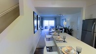 This well-appointed two-floor residence offers a thoughtfully designed interior that combines functionality with contemporary style. The open-plan layout seamlessly connects the dining area to the living room, creating a spacious and cohesive environment ideal for both everyday living and entertaining. The dining area features a sleek white rectangular table surrounded by modern white upholstered chairs, set atop hardwood-style flooring and accented by a decorative framed mirror on the wall. Moving into the living area, plush carpeting adds warmth underfoot, while a track lighting system overhead provides adjustable, even illumination throughout. The living room is comfortably furnished with a sofa and coordinating seating, complemented by a stylish floor lamp and lush indoor greenery that adds a natural touch to the space. A large sliding glass door at the far end of the living area floods the room with natural light and opens onto a private balcony, offering an inviting indoor-outdoor connection. The neutral color palette of soft whites and greys throughout the space creates a calm, airy ambiance. Spanning two floors, this residence provides ample room across multiple levels, making it a versatile and comfortable home.