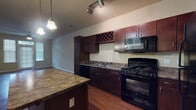 This single-floor residence offers a thoughtfully designed layout that balances functionality and comfort. The kitchen serves as a central highlight, featuring rich dark espresso cabinetry, granite-style countertops, and a full suite of black appliances including a gas range, built-in microwave, refrigerator, and dishwasher. A built-in wine rack adds a distinctive touch, while a large central island provides generous prep and dining space. Pendant lights and track lighting illuminate the kitchen, enhancing its warm ambiance. Warm-toned hardwood flooring flows throughout the main living areas, complementing the neutral walls and creating a cohesive, inviting atmosphere. The open living area is generously sized, offering flexible arrangement possibilities. The front entry features a glass-paneled door flanked by tall sidelight windows, allowing abundant natural light to fill the space. The bedrooms are finished with plush carpeting and include ceiling fans and window treatments, offering a comfortable and well-lit retreat. The bathroom continues the design language of the home with matching dark cabinetry, a granite-style vanity top, a large mirror, and hardwood-style flooring. Overall, the space presents a clean, cohesive aesthetic with practical finishes throughout.