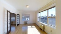 This 2-bedroom, 1-bathroom residence offers a thoughtfully designed single-floor layout that maximizes both comfort and functionality. The living area is bathed in natural light, courtesy of large, multi-pane windows that frame views of the surrounding neighborhood's classic architecture. Warm hardwood floors run throughout, complementing the crisp white walls and elegant crown molding that lend a timeless character to the space. Classic cast-iron radiators provide efficient, even heating and serve as charming architectural accents reflective of the building's heritage. A distinctive French door with divided glass panes adds a refined touch, creating a graceful transition between rooms while maintaining an open, airy feel. The flush-mount ceiling light fixture enhances the room's clean aesthetic. The generous proportions of the main living area allow for flexible furniture arrangements, making it well-suited for both relaxing and entertaining. With its combination of classic detailing, abundant natural light, and hardwood flooring, this residence presents a warm and inviting atmosphere that balances traditional charm with practical everyday living.