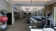 This modern fitness center is designed to provide a comprehensive workout environment within a single, open-plan floor. The space is thoughtfully laid out, with a row of cardio machines — including treadmills and ellipticals — positioned along one side, offering ample room to move freely between equipment. A dedicated strength and free weights area is situated toward the back, stocked with a variety of machines and dumbbells to support a full range of training routines.

The interior design features a neutral color palette with striped carpeted flooring that adds texture while defining workout zones. The ceiling is a standout architectural element, with recessed rectangular coffers framing ceiling fans that promote consistent air circulation throughout the space. Linear LED lighting fixtures provide bright, even illumination, complemented by large windows that allow generous natural light to fill the room and offer views to the outside.

Wall-mounted televisions are positioned strategically for visibility across the floor. The overall ambiance is clean, airy, and professional — striking a balance between functionality and comfort that encourages regular use. Entry doors with glass panels connect the fitness center to adjacent areas, enhancing its accessibility and integration within the broader facility.