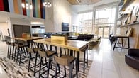 This amenity space is designed with a contemporary aesthetic and offers a diverse range of facilities across a single level. The entrance lobby features polished tile flooring, a textured wave-panel accent wall, exposed brick detailing, and elevator access, with potted greenery and a patterned area rug adding warmth to the clean, modern palette.

The main lounge area is a standout, boasting soaring ceilings, a dramatic floor-to-ceiling marble accent wall, a wall-mounted television, and a linear gas fireplace. Seating includes deep-toned sofas, gold-framed armchairs, and a marble coffee table, all oriented toward expansive floor-to-ceiling windows that frame views of the surrounding urban streetscape. Adjacent to this is a sun-drenched café-style seating area furnished with round tables and vibrant yellow chairs, bathed in natural light from the glass curtain wall.

The game and social lounge offers a billiards table, shuffleboard, comfortable sectional seating in navy and grey tones, a bar-height counter, and a linear fireplace, creating an engaging social environment. A live-edge communal table with bar stools provides additional gathering space nearby.

The fitness center is extensively equipped with rows of treadmills, ellipticals, stair climbers, stationary bikes, and strength machines, complemented by a dedicated free-weights and functional training studio with mirrored walls and a large display screen.
