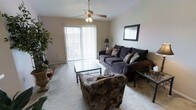 This single-floor residential space offers a comfortable and well-lit layout designed for everyday living. The living room serves as the heart of the home, featuring plush wall-to-wall carpeting that adds warmth underfoot. A ceiling fan with a light fixture is centrally mounted, providing both air circulation and ambient lighting. Large vertical blinds frame a sliding glass door that fills the room with natural light and offers a view to the outdoors, creating an open, airy atmosphere. The textured ceiling adds a classic touch to the overall aesthetic. The living area provides ample space for seating arrangements, making it well-suited for both relaxing and entertaining. Neutral wall tones throughout keep the space feeling clean and versatile, allowing for a variety of décor styles. The single-floor layout ensures seamless flow and easy accessibility across the entire living space. Overall, the residence combines functional design with comfortable living, highlighted by its inviting natural light, practical ceiling fan, and easy indoor-outdoor connection through the sliding glass door.