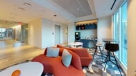 This thoughtfully designed single-floor amenity space combines multiple functional zones to support a variety of work and social needs. The layout flows seamlessly from one area to the next, unified by a contemporary design palette of warm wood tones, bold accent colors, and mixed flooring materials including light hardwood and geometric patterned tile.

A glass-enclosed meeting room anchors one end of the space, furnished with a round white table and teal upholstered chairs. Floor-to-ceiling windows on two sides flood the room with natural light while offering views of surrounding buildings and greenery, creating an inviting atmosphere for small group discussions.

Adjacent to the meeting room, a coworking zone provides individual workstations equipped with desktop computers, mounted against neutral wall panels with vertical sconce lighting. A slatted wood partition elegantly separates this area from the lounge without fully enclosing it.

The central lounge area features a bold terracotta sectional sofa and accent seating arranged around small round tables, encouraging casual conversation. Nearby, a well-appointed refreshment bar with navy cabinetry, a coffee machine, and counter seating rounds out the space, offering a functional hub for breaks and informal gatherings. Recessed and cove lighting throughout contributes to the space's warm, polished ambiance.