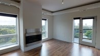 This single-floor space presents a clean, contemporary design with an open living area that combines style and functionality. The room is anchored by a wall-mounted linear fireplace with a stainless steel surround, serving as a striking focal point against the crisp white walls. Warm-toned hardwood flooring extends throughout, adding a natural contrast to the modern aesthetic.

Natural light flows generously into the space through multiple windows, each framing views of lush, tree-lined surroundings and a nearby urban skyline, creating a pleasant connection to the outdoors. A sliding glass door or large window on the right side of the room further enhances this openness, potentially leading to an outdoor area.

The ceiling features a subtle tray detail with integrated track lighting, providing both ambient illumination and an architectural design element that adds depth to the room. Neutral tones dominate the palette throughout, creating a versatile backdrop well-suited for a variety of interior styles. The overall ambiance is calm, airy, and refined, reflecting a thoughtfully designed modern living environment.