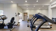 Located at 125 W Olive Ave in Monrovia, California, this fitness center offers a well-equipped workout space designed to support a range of exercise routines. The single-floor layout is efficiently organized, making good use of its footprint to accommodate multiple exercise stations without feeling cramped.

The space features recessed ceiling lighting throughout, providing consistent, even illumination across the entire room. Structural columns are integrated into the layout and contribute to the room's defined zones. The flooring is a durable speckled carpet-style material well-suited for fitness use, and dark wood baseboards add a clean finish along the walls.

Cardio equipment is a highlight of this fitness center, with multiple treadmills — including True and 7000 Series models — positioned along the center of the room. Cycling options include both upright and recumbent stationary bikes. An elliptical trainer and a rowing-style machine round out the cardio offerings. A weight bench and a cable/pulley strength machine are also present, supporting resistance and strength training.

Natural light enters through large windows and a sliding glass door on one side of the room, offering views of the outdoor surroundings and creating an open, airy atmosphere. A wall-mounted sanitizing dispenser and a security camera are also present. The space includes a dedicated exit door, ensuring convenient and safe access.
